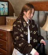 Person wearing a brown floral cardigan in a room with wooden furniture and a lamp.